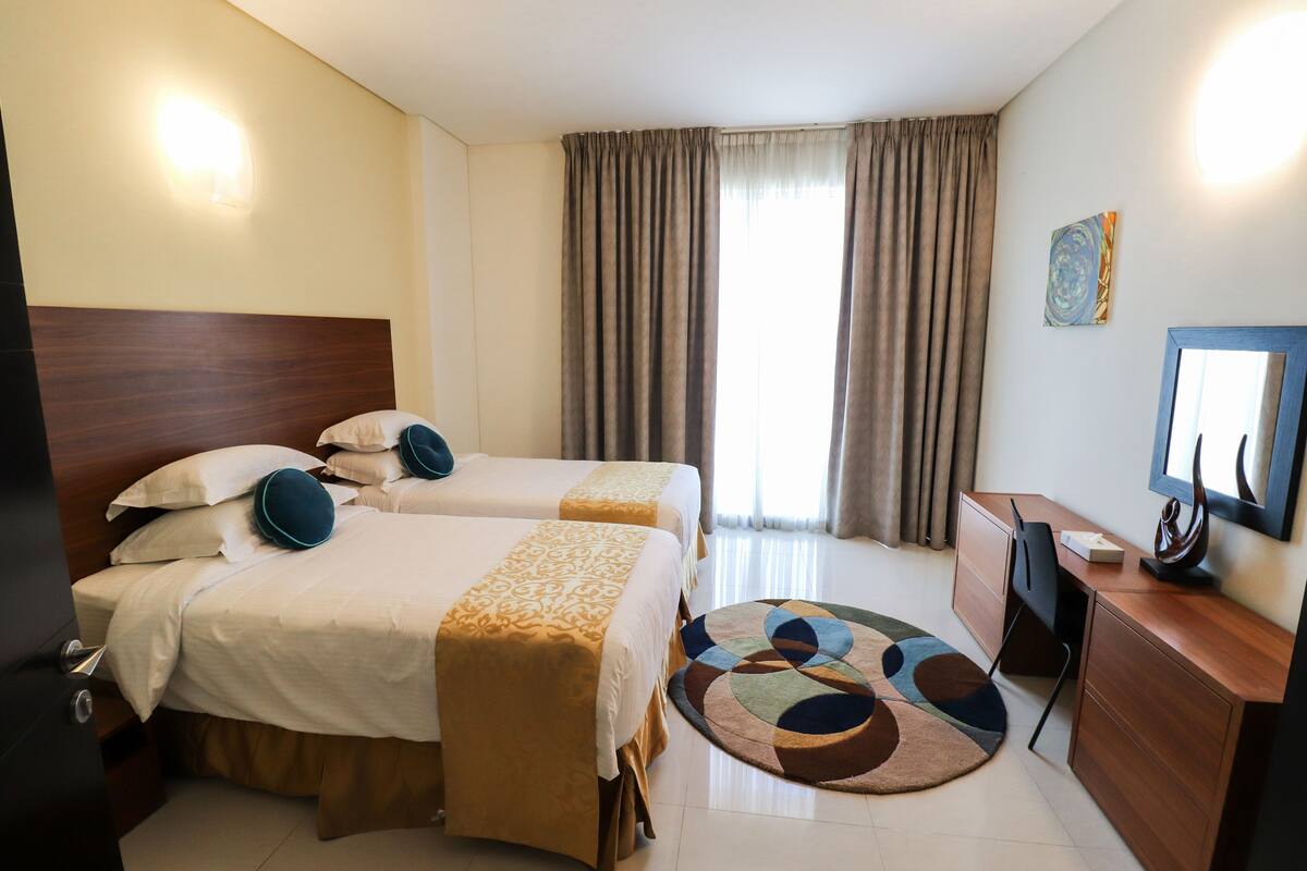 A hotel room with two beds, a desk, a chair, a TV, and a window with curtains.