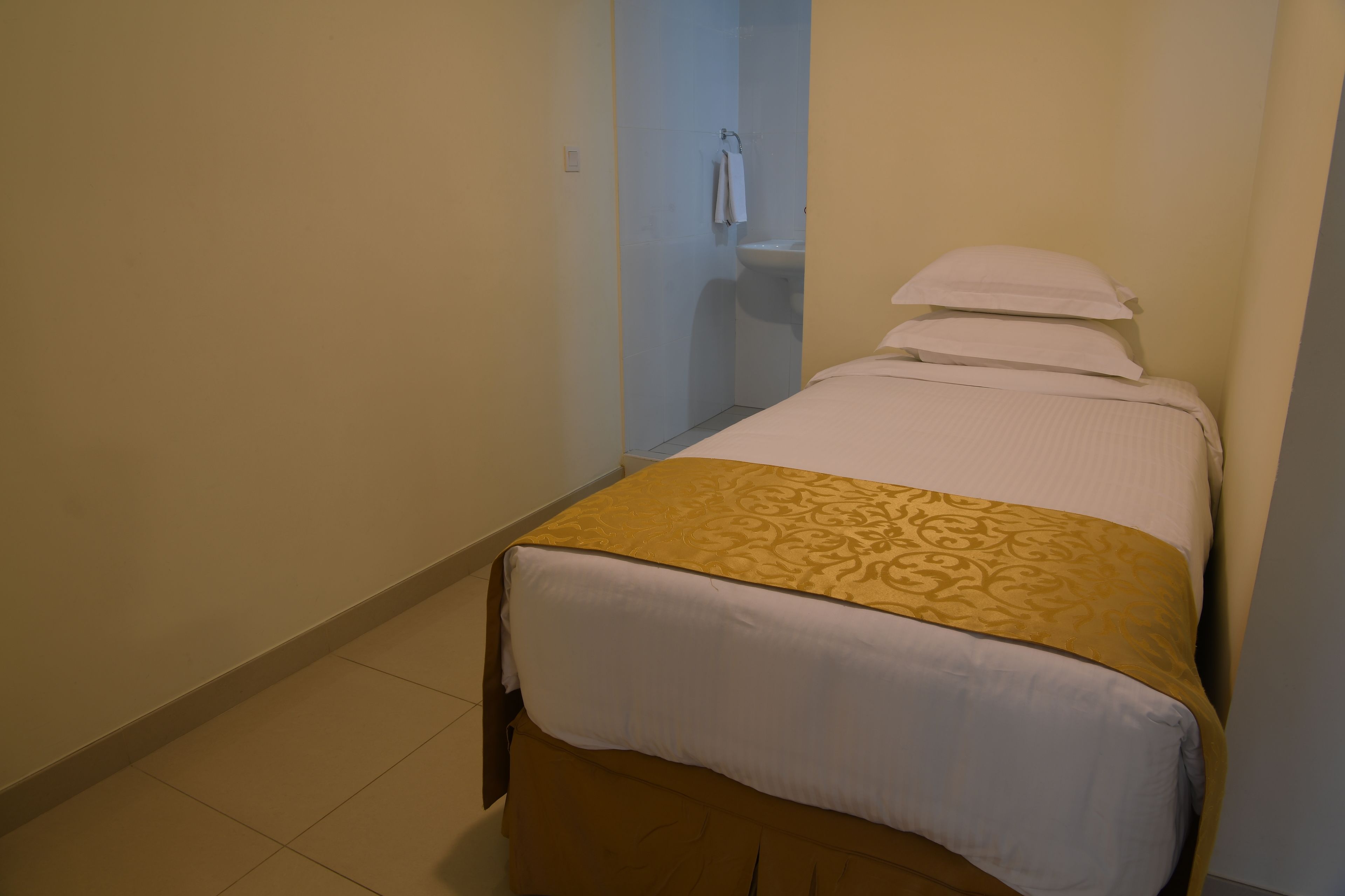 A small, single-bed room with a beige floor, a white bedspread, and a gold-patterned blanket.