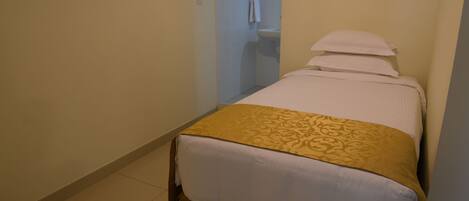 A small, single-bed room with a beige floor, a white bedspread, and a gold-patterned blanket.