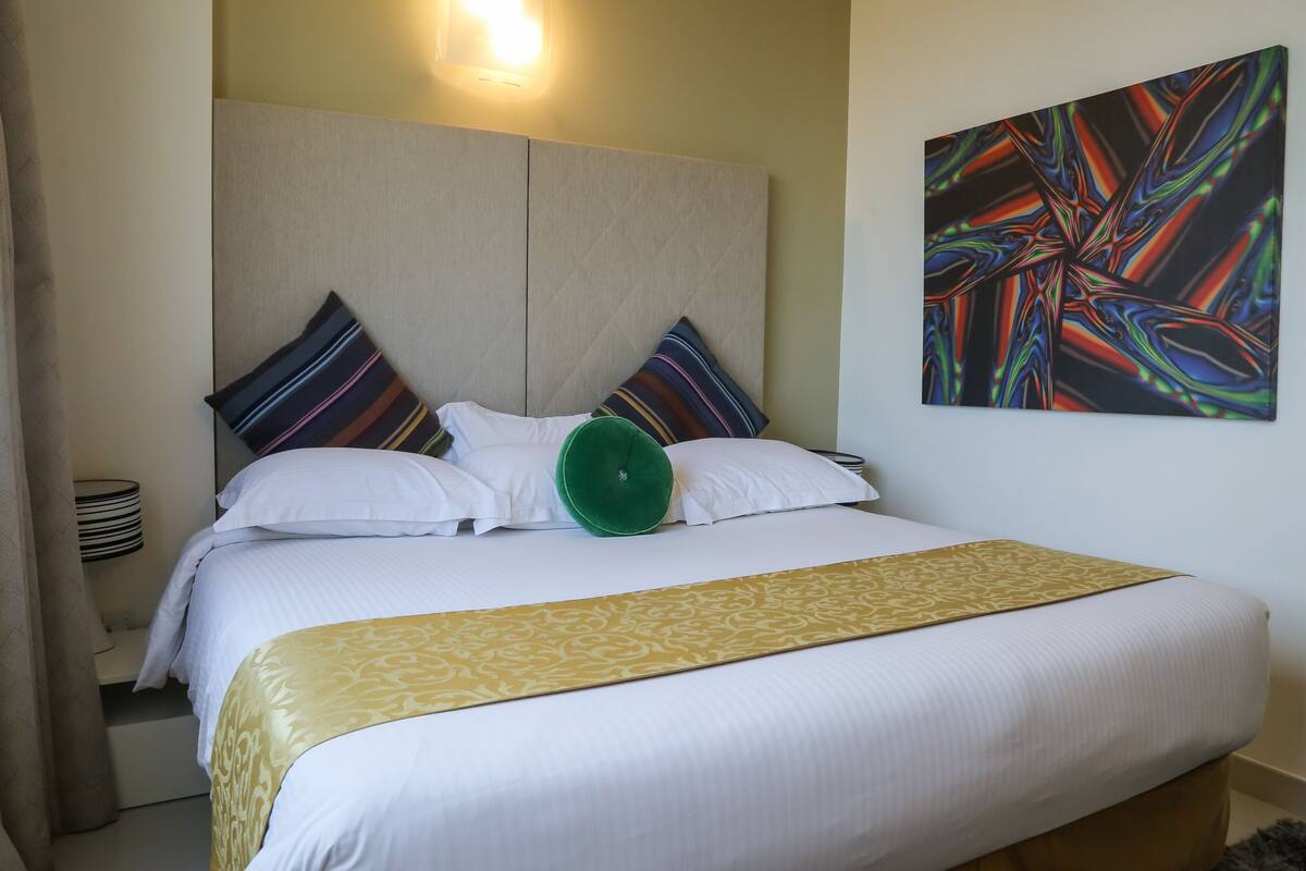 A hotel room with a large bed, a green accent pillow, and a colorful abstract painting on the wall.