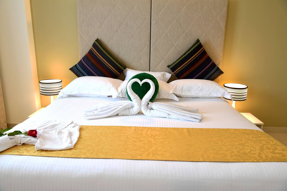 A neatly made bed with towels arranged in a heart shape, a rose, and a decorative throw.