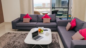 A modern living room with a grey sofa, a white coffee table, and a laptop. There is a view of a cityscape through a large window. - Gulf Suites Hotel Amwaj (Amwaj Islands)