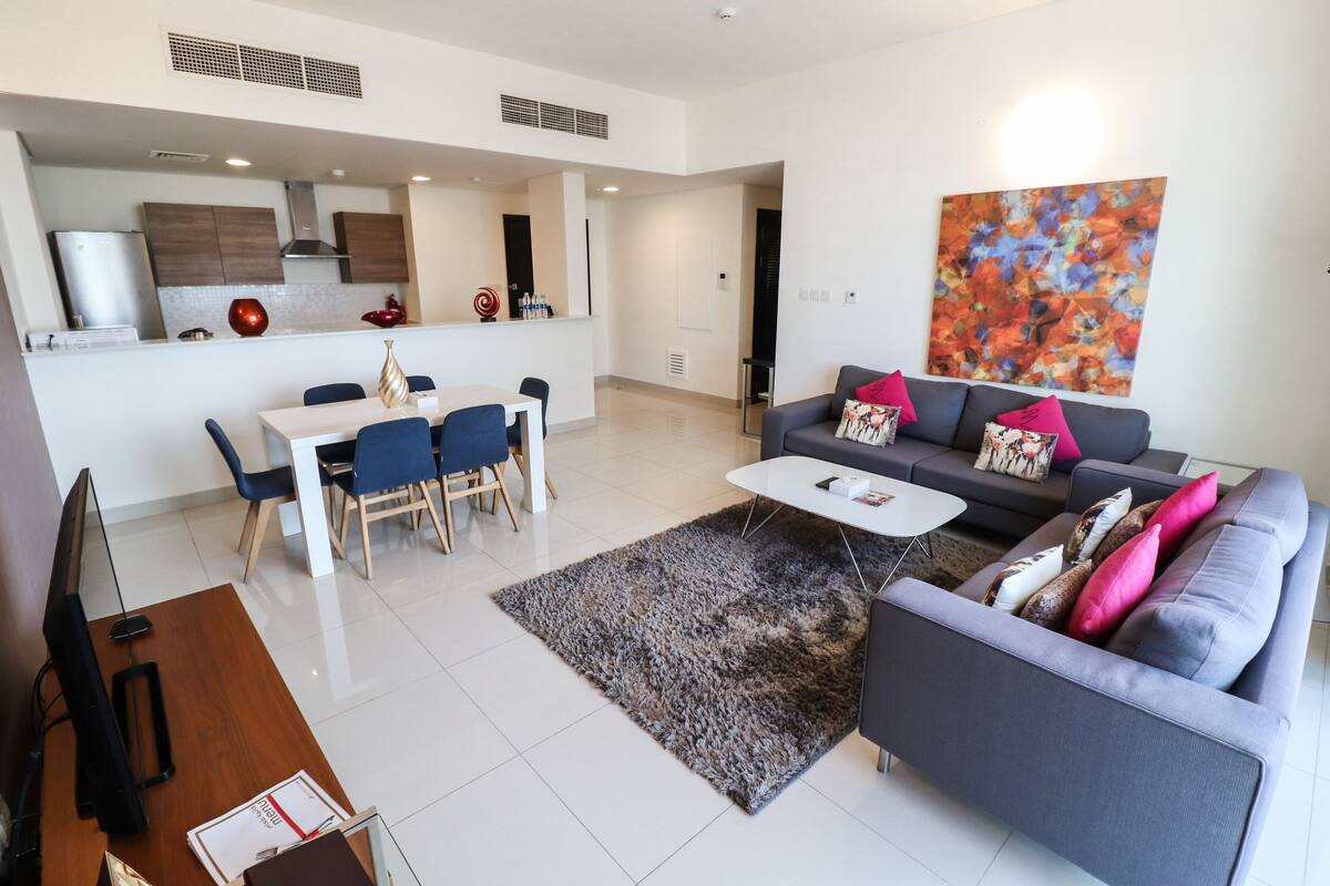 A modern living room with a grey sofa, a coffee table, and a dining area with a white table and blue chairs.