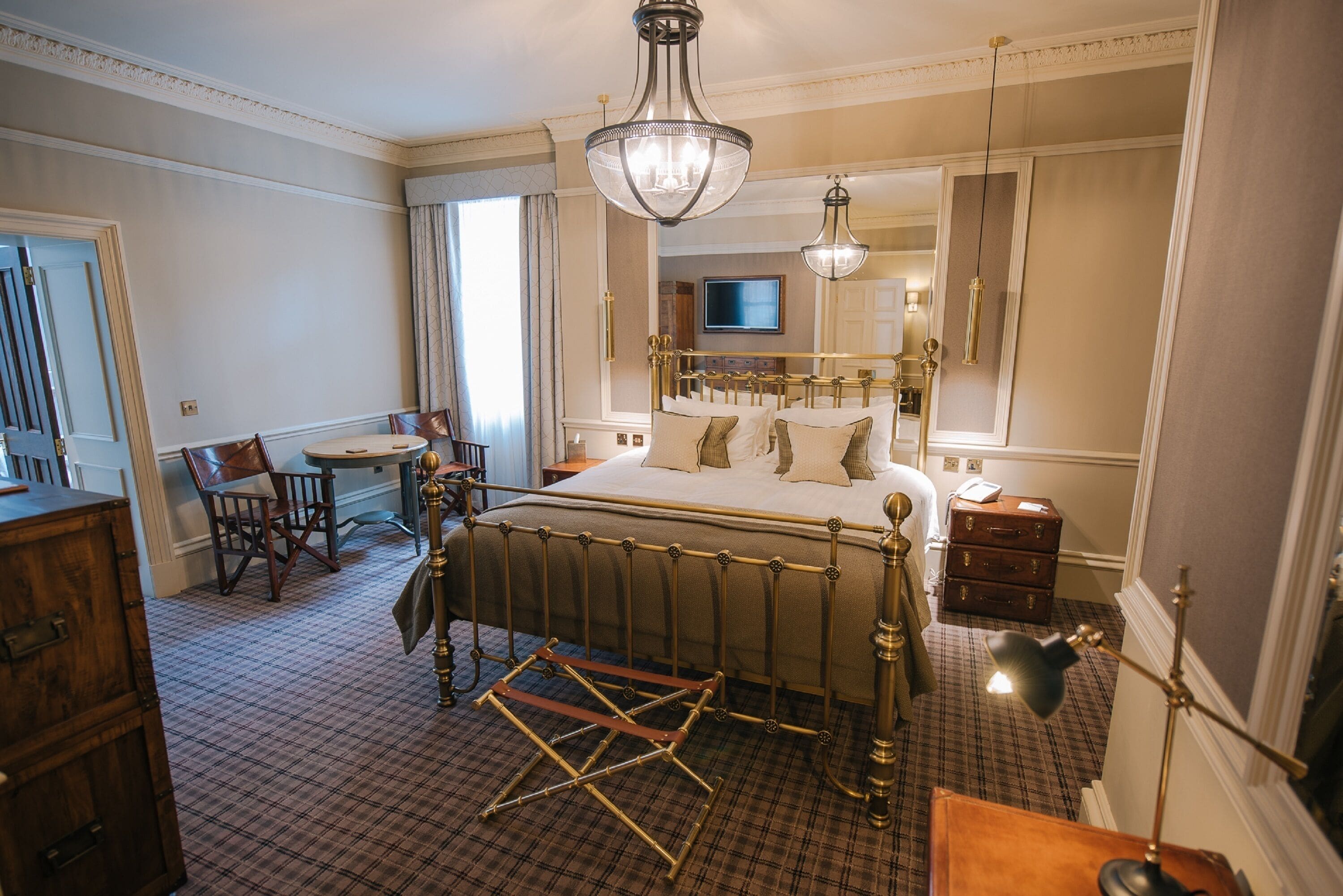 A hotel room with a large bed, a desk, a chair, a dresser, a suitcase, and a television.
