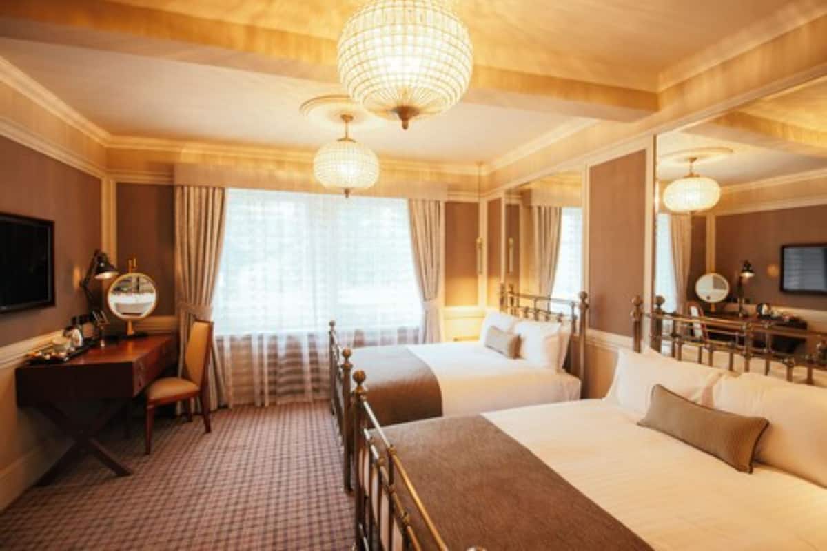 A hotel room with two beds, a desk with a chair, a large mirror, and a chandelier.