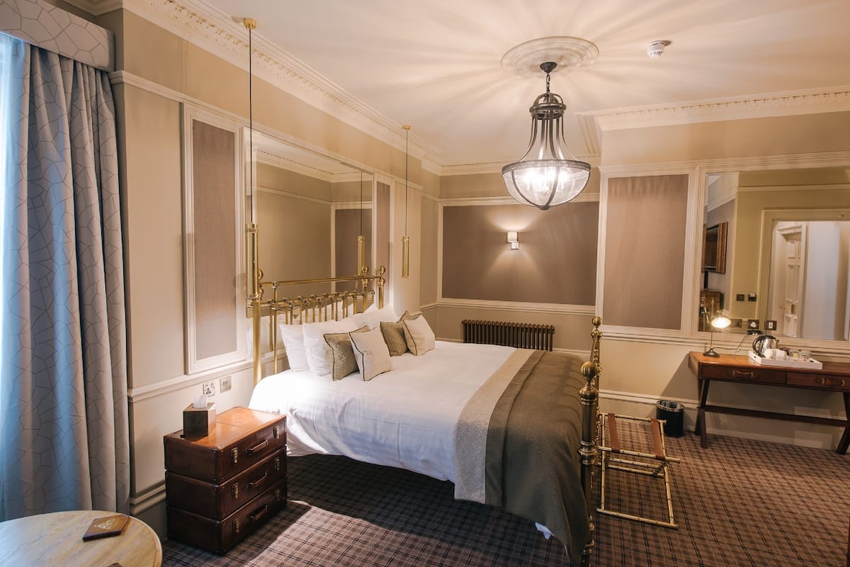 A hotel room with a large bed, a chest of drawers, a desk, a mirror, and a hanging light fixture.