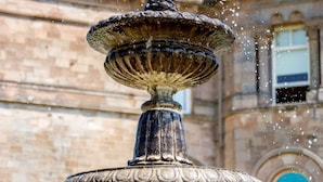 Una fuente de piedra de varios niveles con agua brotando del nivel superior, frente a un edificio histórico con ventanas de arco y fachada de ladrillo.
