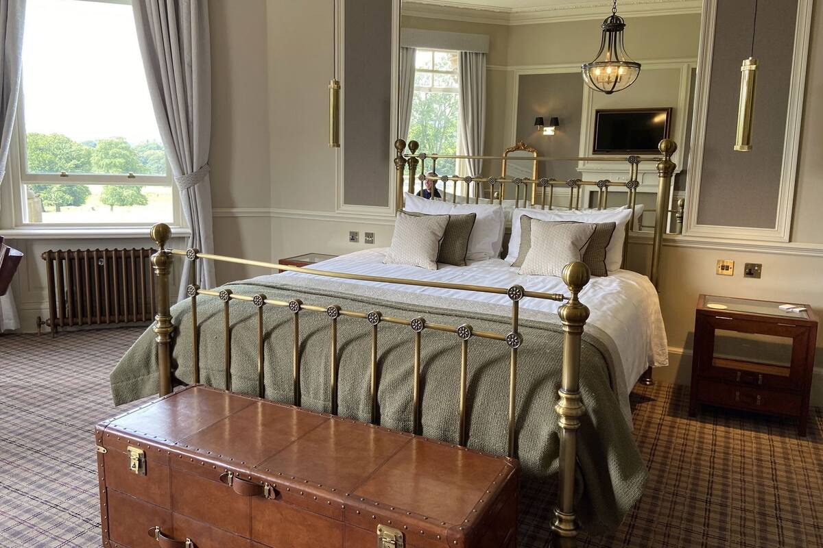 A bedroom with a four-poster bed, a wooden chest, a radiator, a television, and a window with curtains.