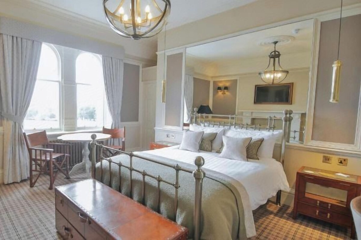 Una habitación de hotel con una cama, un escritorio, una silla, un aparador, un espejo y un ventanal con cortinas.