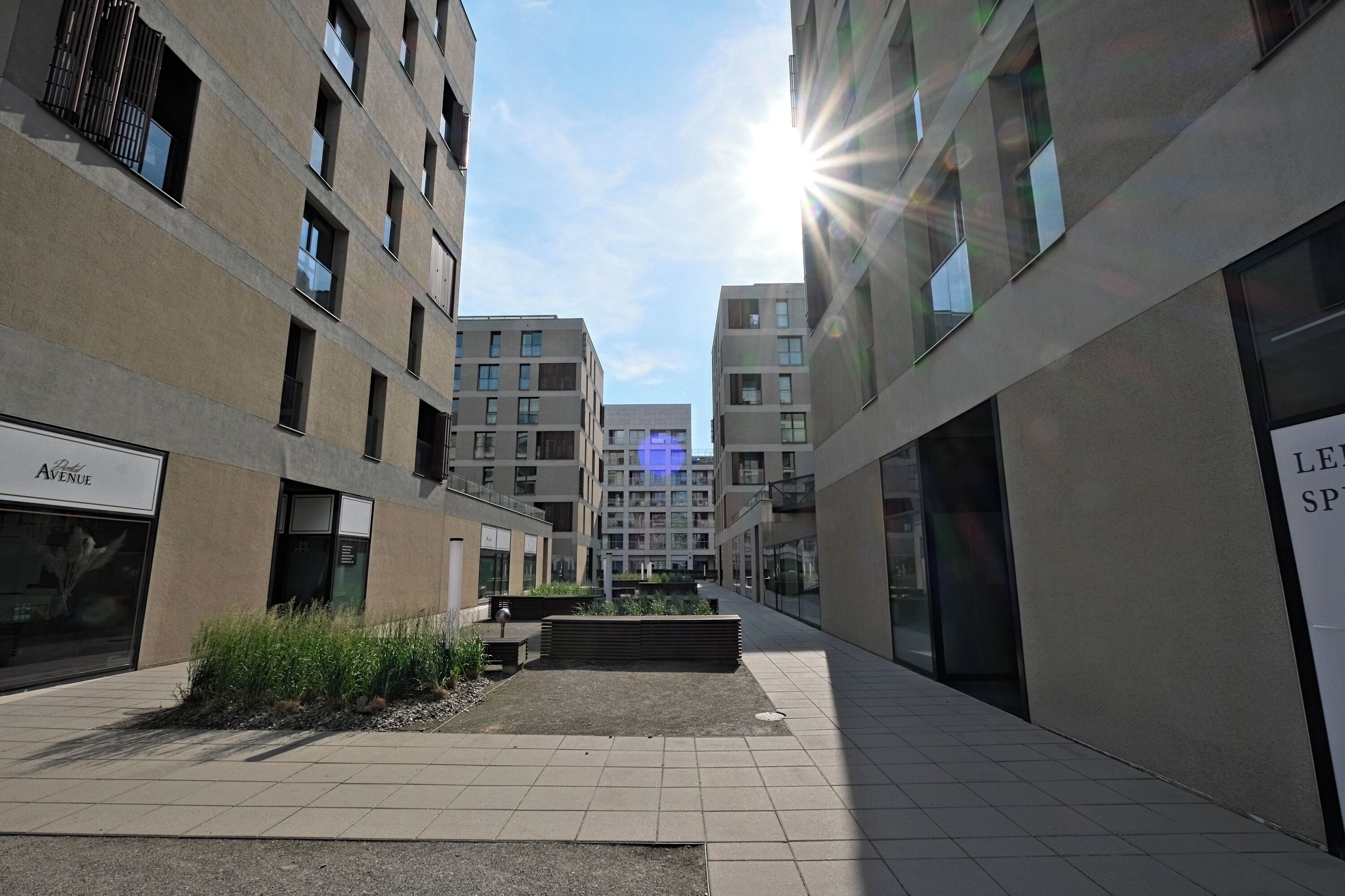 Un patio urbano moderno flanqueado por edificios, con un jardín central y cielo despejado.