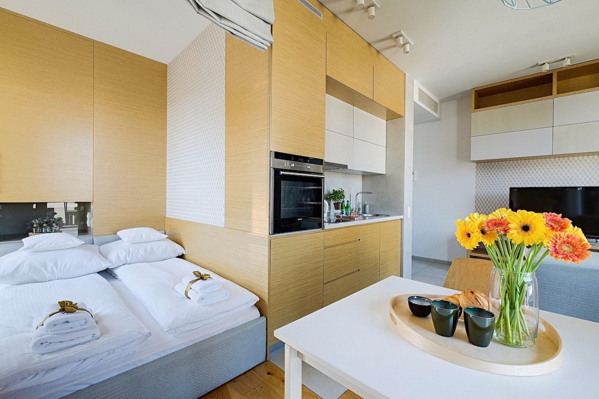 Quarto de hotel moderno com cama, pequeno espaço de cozinha, área de refeições e um vaso de flores.