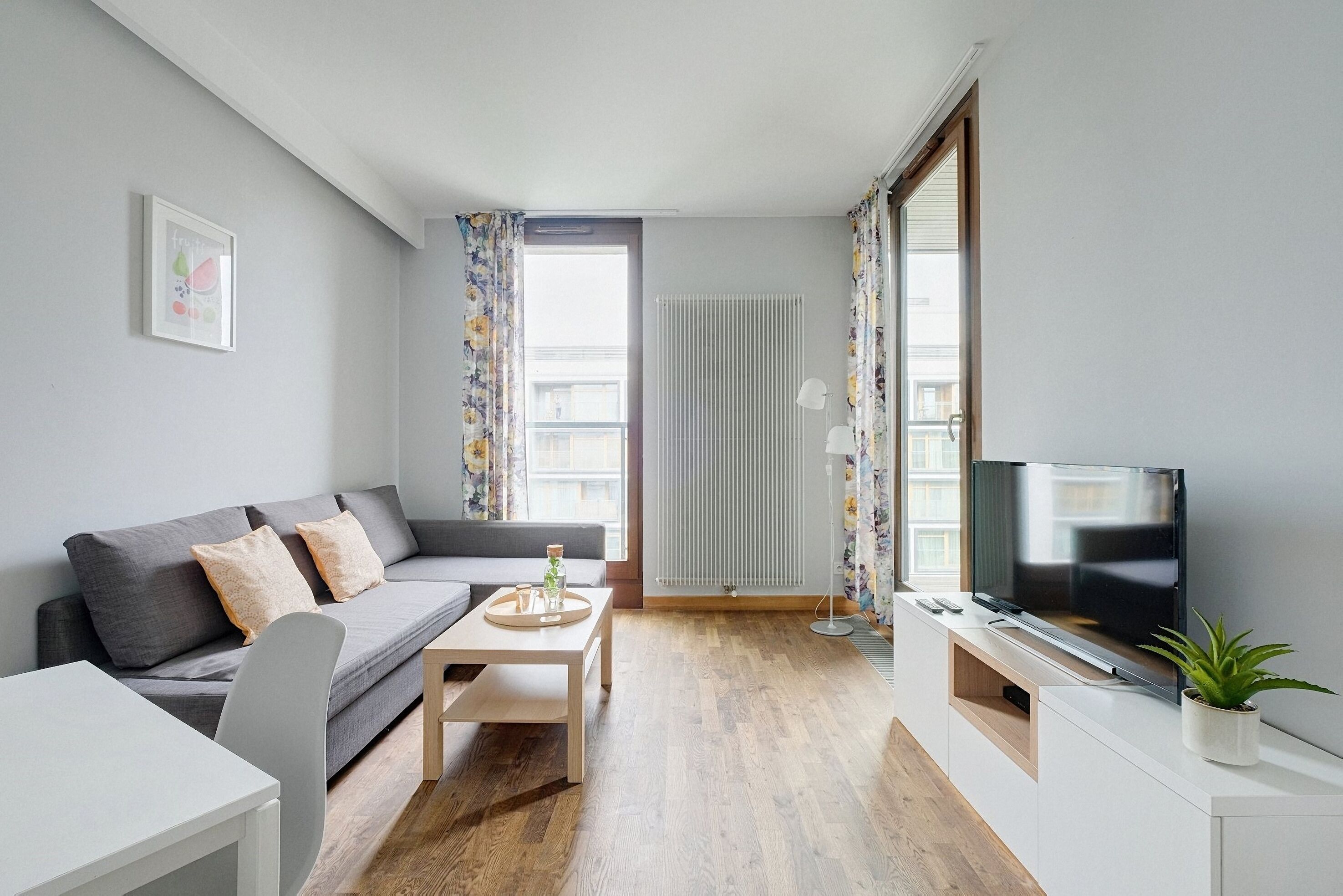 A modern living room with a grey sofa, a wooden coffee table, a TV on a white cabinet, and a potted plant.