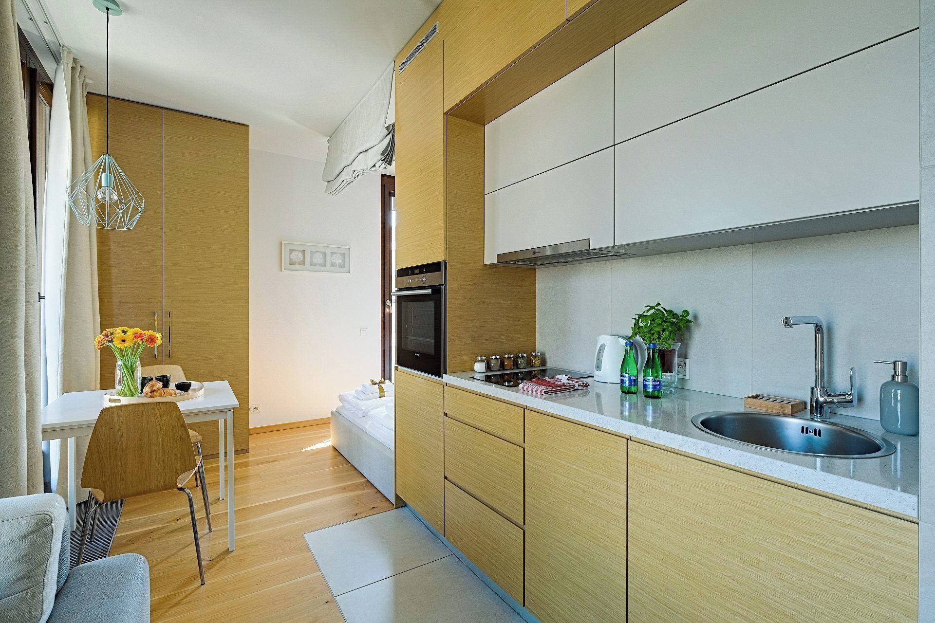 Una cocina moderna con armarios de madera, un lavamanos de acero inoxidable y un horno empotrado. Hay una zona de comedor con una mesa y sillas, y un florero con flores sobre la mesa.