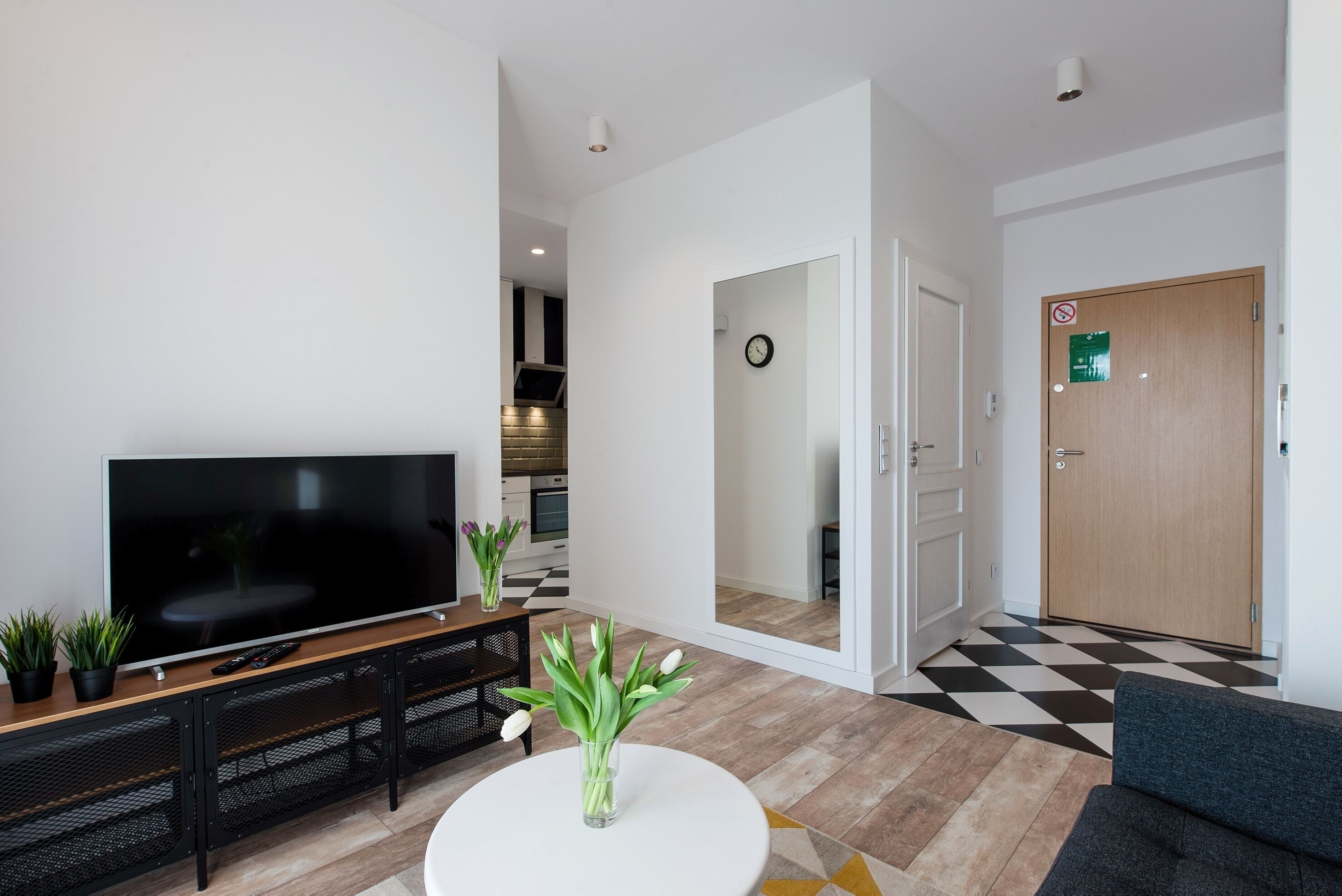 Ein modernes Wohnzimmer mit Flachbildfernseher, rundem Couchtisch und einer Couch. Ein Flur führt zur Küche, und eine Tür trägt ein grünes Schild.