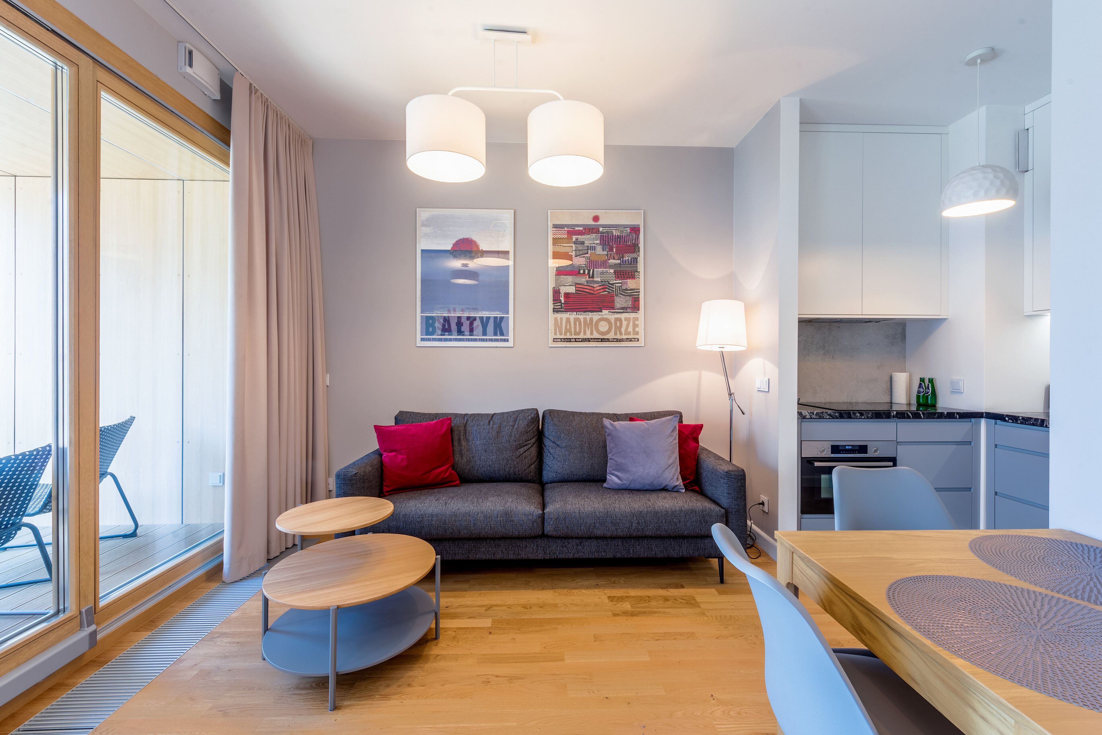 Ett modernt vardagsrum med en grå soffa, röda och blå kuddar, ett träbord och ett matbord med en bordsduk. På väggen hänger två affischer. Det finns också ett köksområde med en spis och skåp, samt ett stort fönster med gardiner.