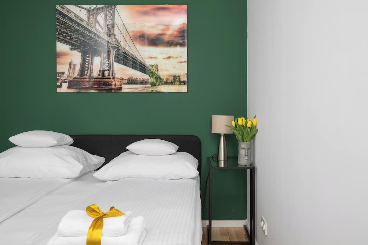 Quarto de hotel com cama, almofadas brancas, cama bem arrumada com toalha amarela, mesa de cabeceira com abajur e um vaso de flores amarelas, e uma grande pintura de paisagem urbana na parede.
