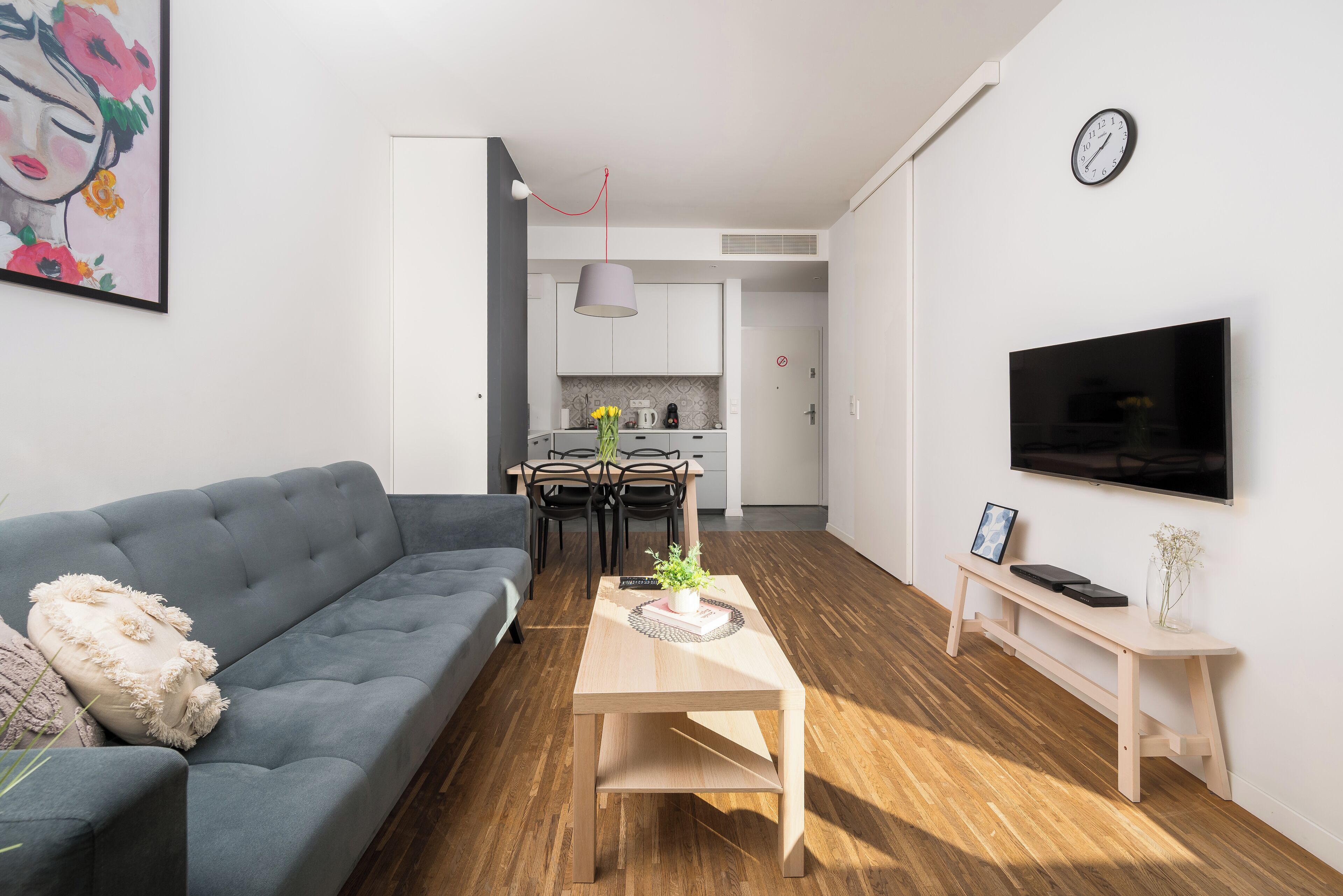 Un salon moderne avec un canapé gris, une table basse en bois, un téléviseur fixé au mur et une cuisine en arrière-plan.
