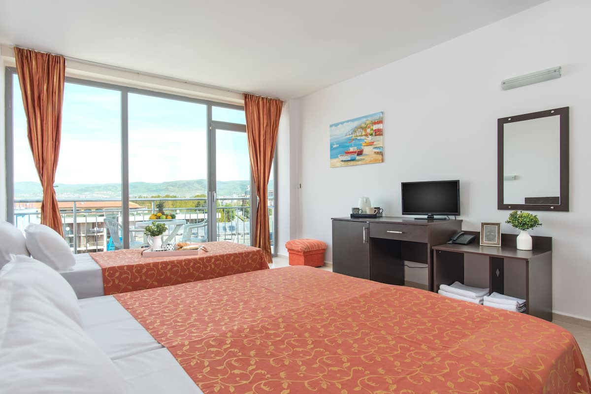 A hotel room with two beds, a desk with a TV, a window with curtains, a painting, and a view of the city.