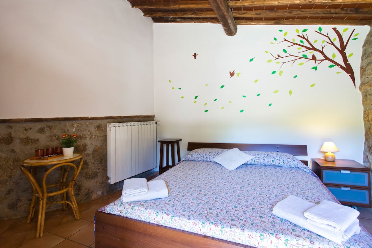 Een slaapkamer met een bed, een houten hoofdbord, een nachtkastje met een lamp, een bijzettafeltje met een plant en een muur met een boomdecoratie.