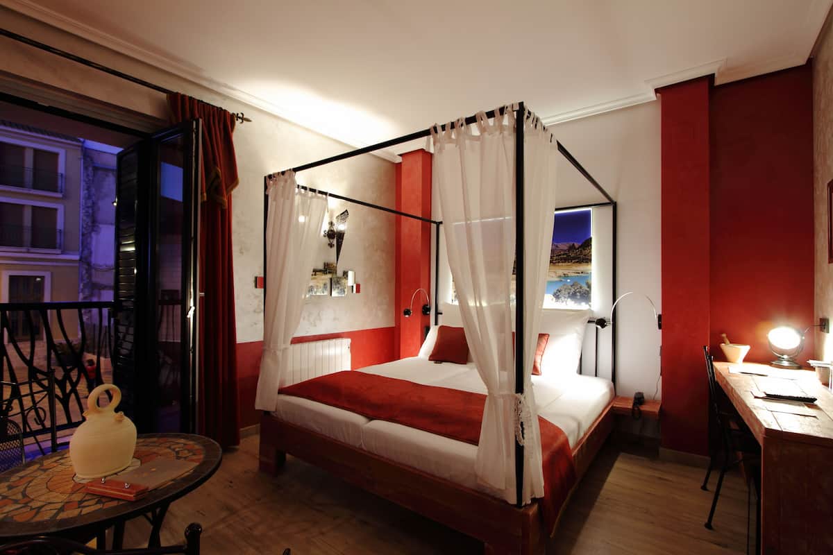 A four-poster bed with a canopy, a desk with a lamp, and a view of the outdoors through a window.