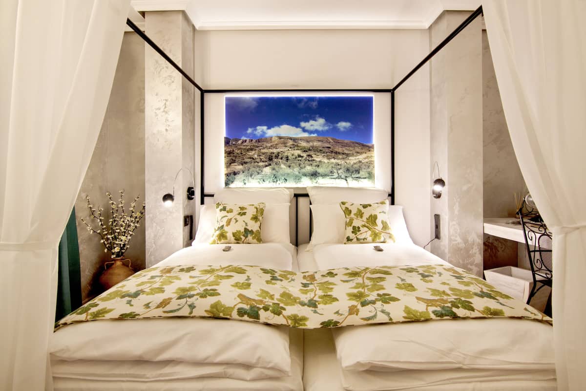 A bedroom with a bed centered under a large window, a vase with branches, and a small table with a chair.