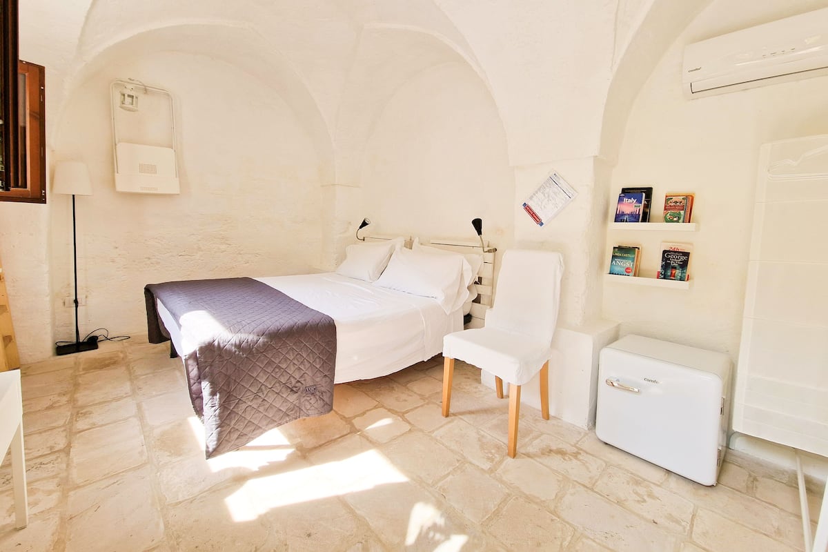 A bedroom with a bed, a chair, a small table, a radiator, and a shelf with books.