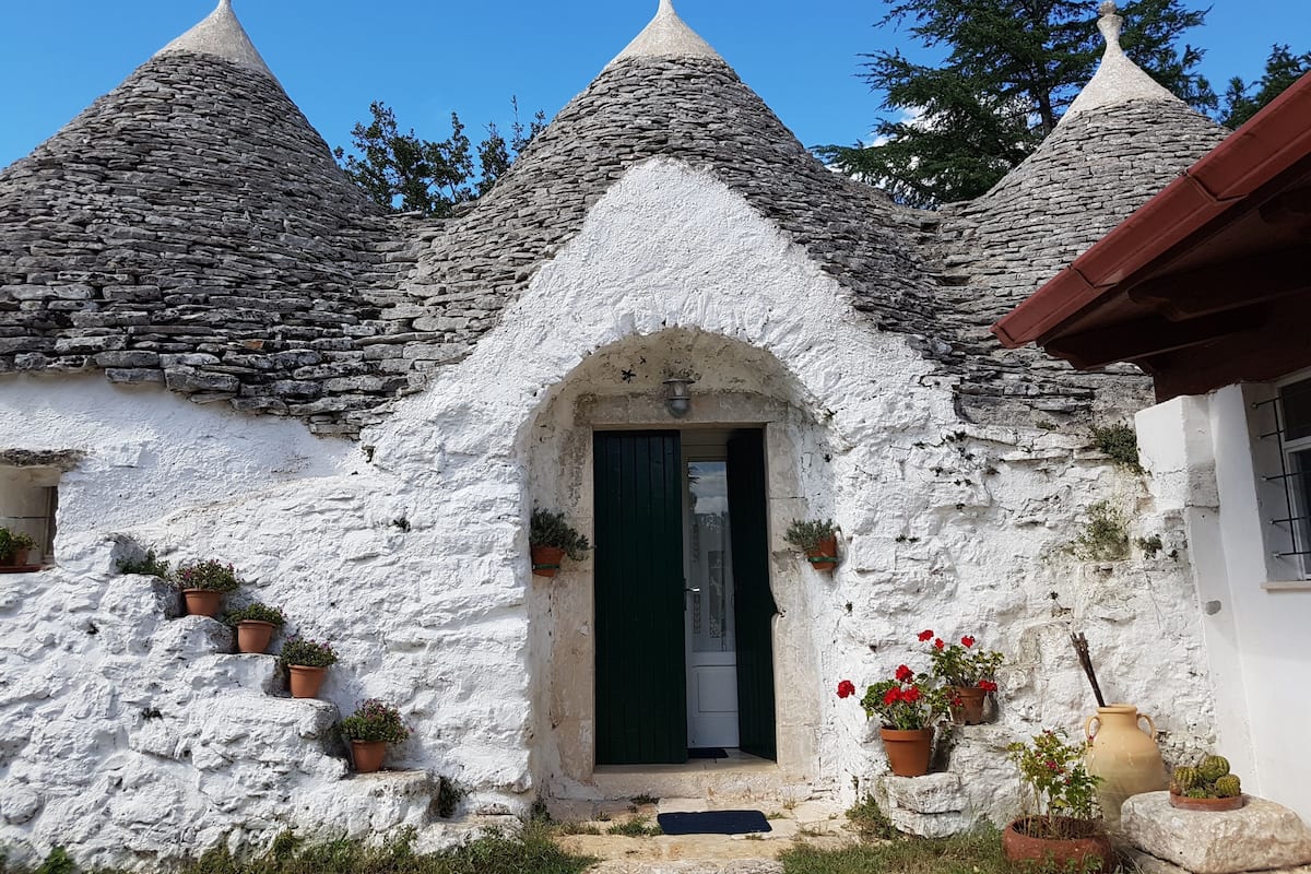 A traditional stone house with a white exterior, a green door, and multiple potted plants around the entrance.