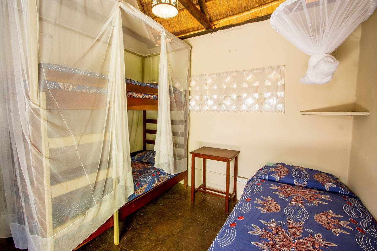Una habitación con dos camas, una con mosquitero, una mesita de madera y un estante en la pared.