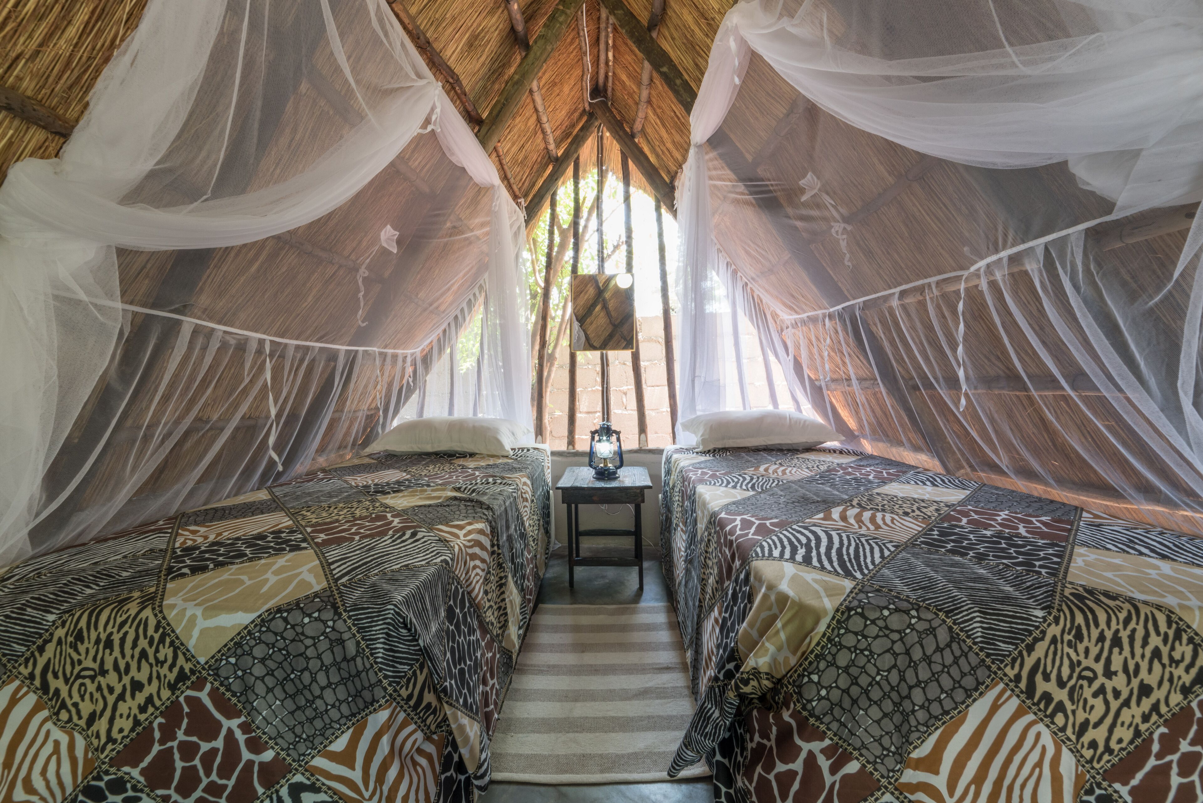 A room with two beds, each with patterned bedding, a small table with a lantern, and a thatched roof.