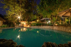 A nighttime pool area with stone walls, trees, and outdoor seating under umbrellas. - Victoria Falls Backpackers (Victoria Falls)