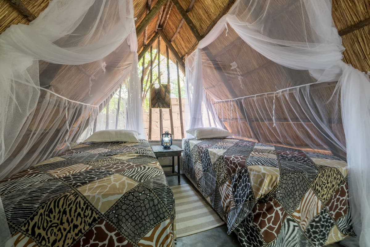 Una habitación con dos camas, cada una con ropa de cama estampada, una mesita con una lámpara y un mosquitero colgando.