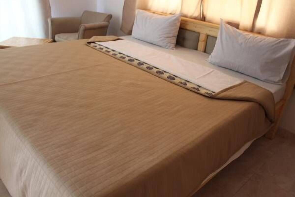 A neatly made bed with a beige bedspread and a decorative runner, a wooden headboard, and a chair in the background.