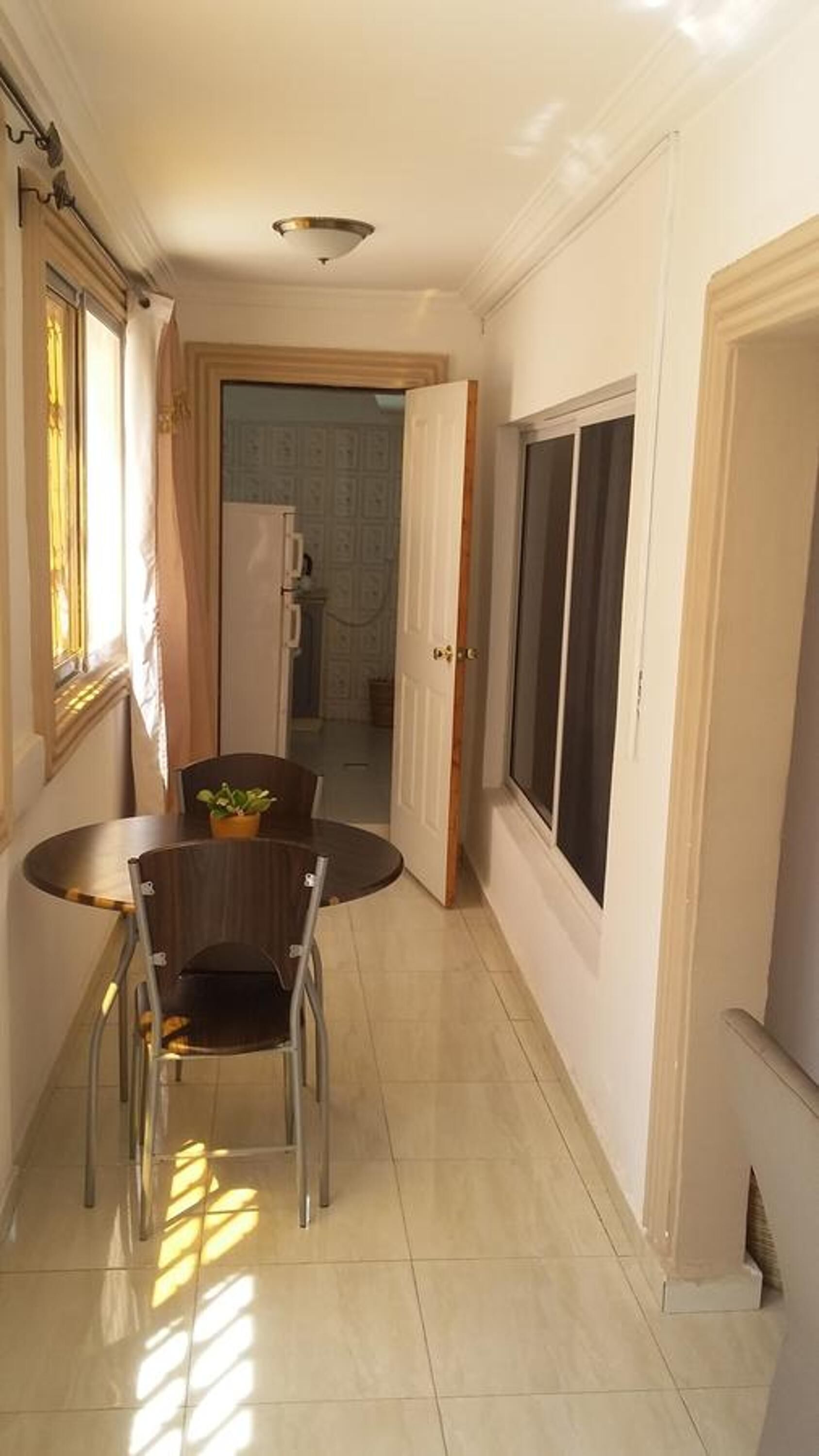 A narrow hallway with a dining table and chairs, a large window, and a door leading to another room.