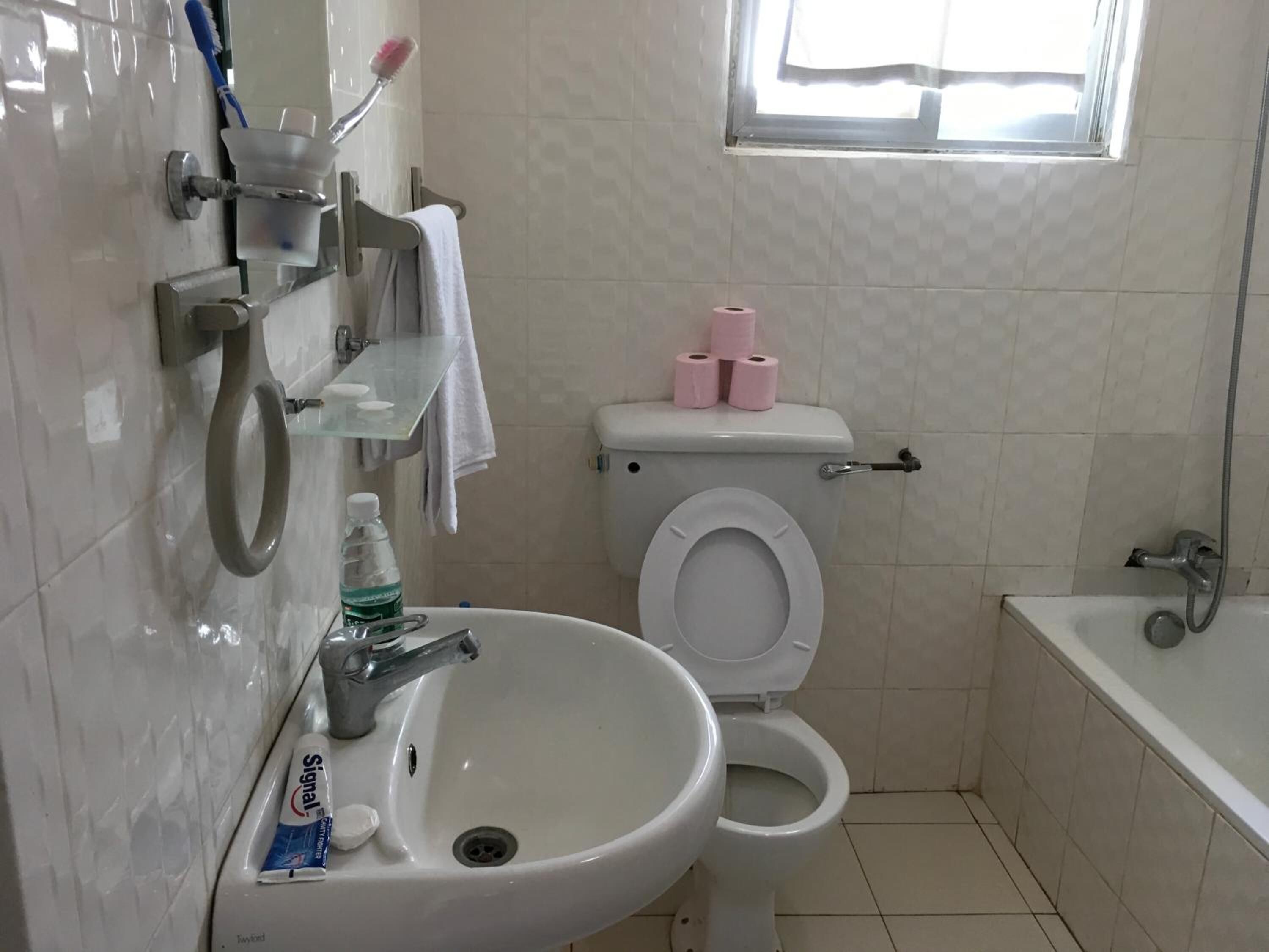 Une salle de bain avec un lavabo, des toilettes et une baignoire, tous de couleur blanche. Il y a une fenêtre au-dessus des toilettes, et une étagère avec une brosse à dents et du dentifrice.