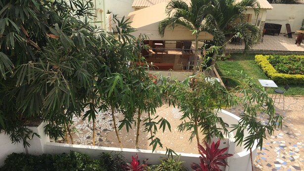 Uma área de pátio externa com piso ladrilhado, vários vasos de plantas e cadeiras dobráveis brancas.