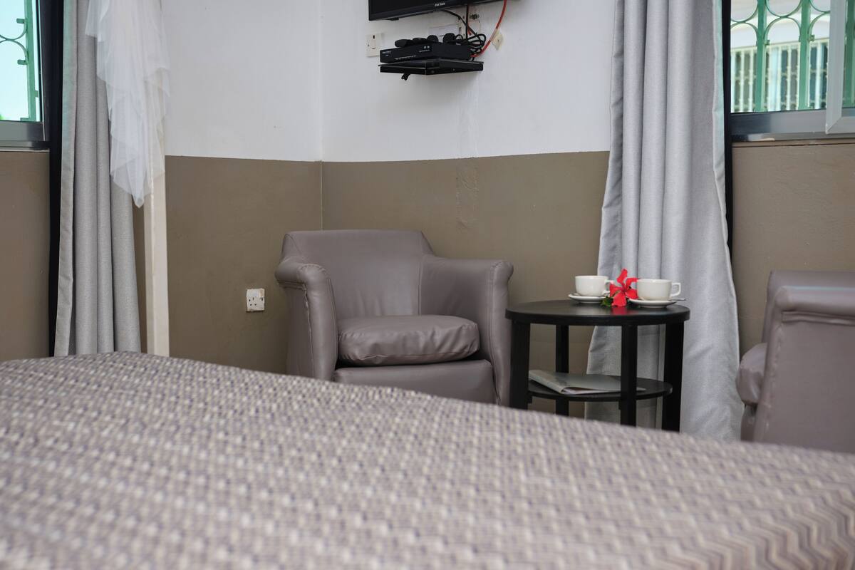A hotel room with a bed, a grey armchair, a small table with a cup and a red flower, and a TV mounted on the wall.