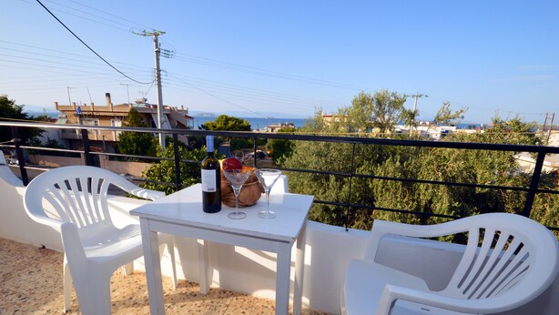A balcony with a table, two white plastic chairs, a bottle of wine, and a martini glass. - Koukounari Rooms (Agistri)