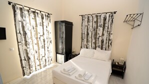 A hotel room with a bed, curtains, a TV, a small fridge, a bedside table, and a nightstand. - Koukounari Rooms (Agistri)