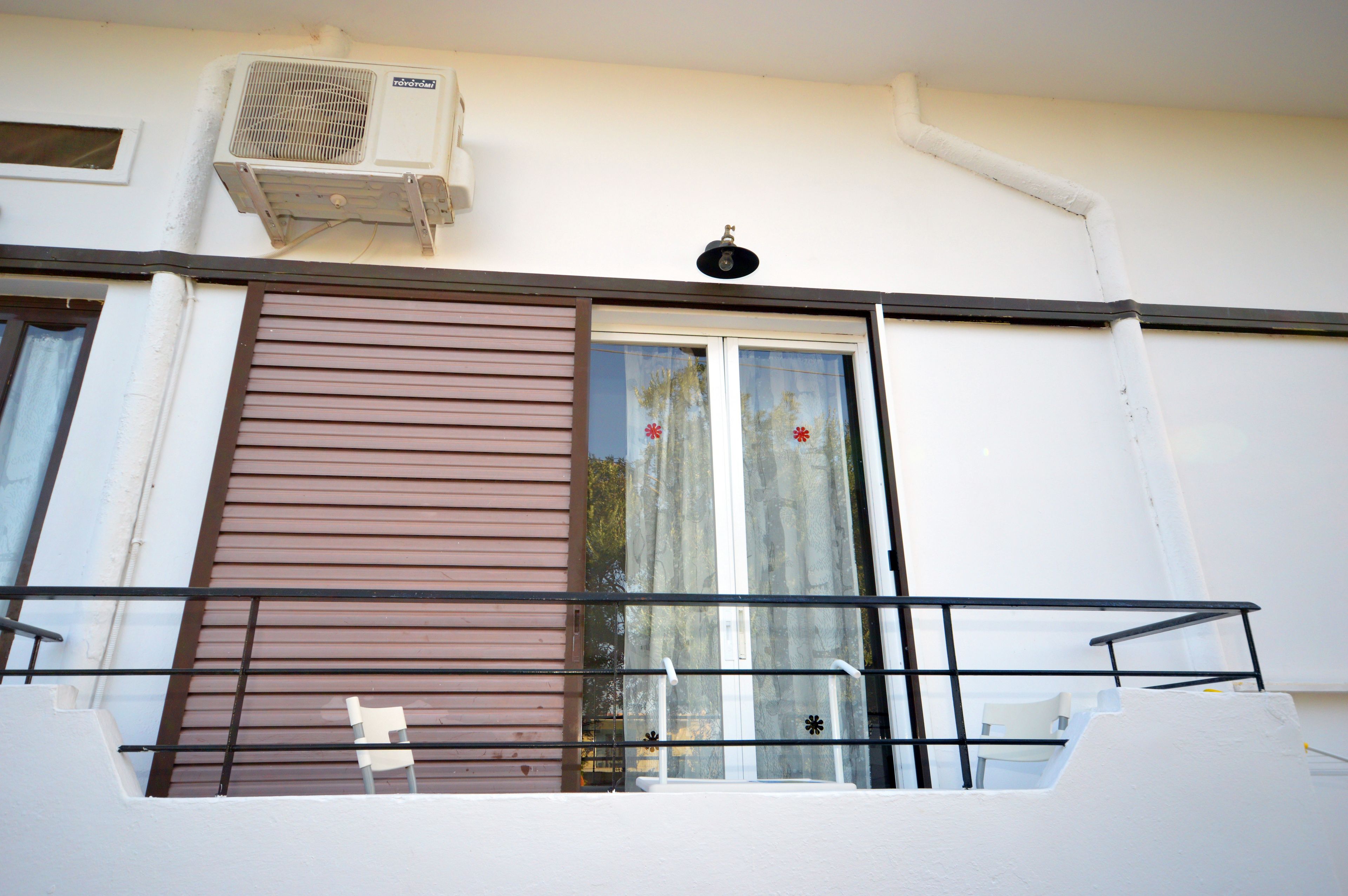 A balcony with a metal railing, a window with a brown shutter, and an air conditioning unit mounted on the wall.