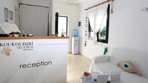A reception area with a desk, a water dispenser, a white sofa, and a window with curtains. - Koukounari Rooms (Agistri)
