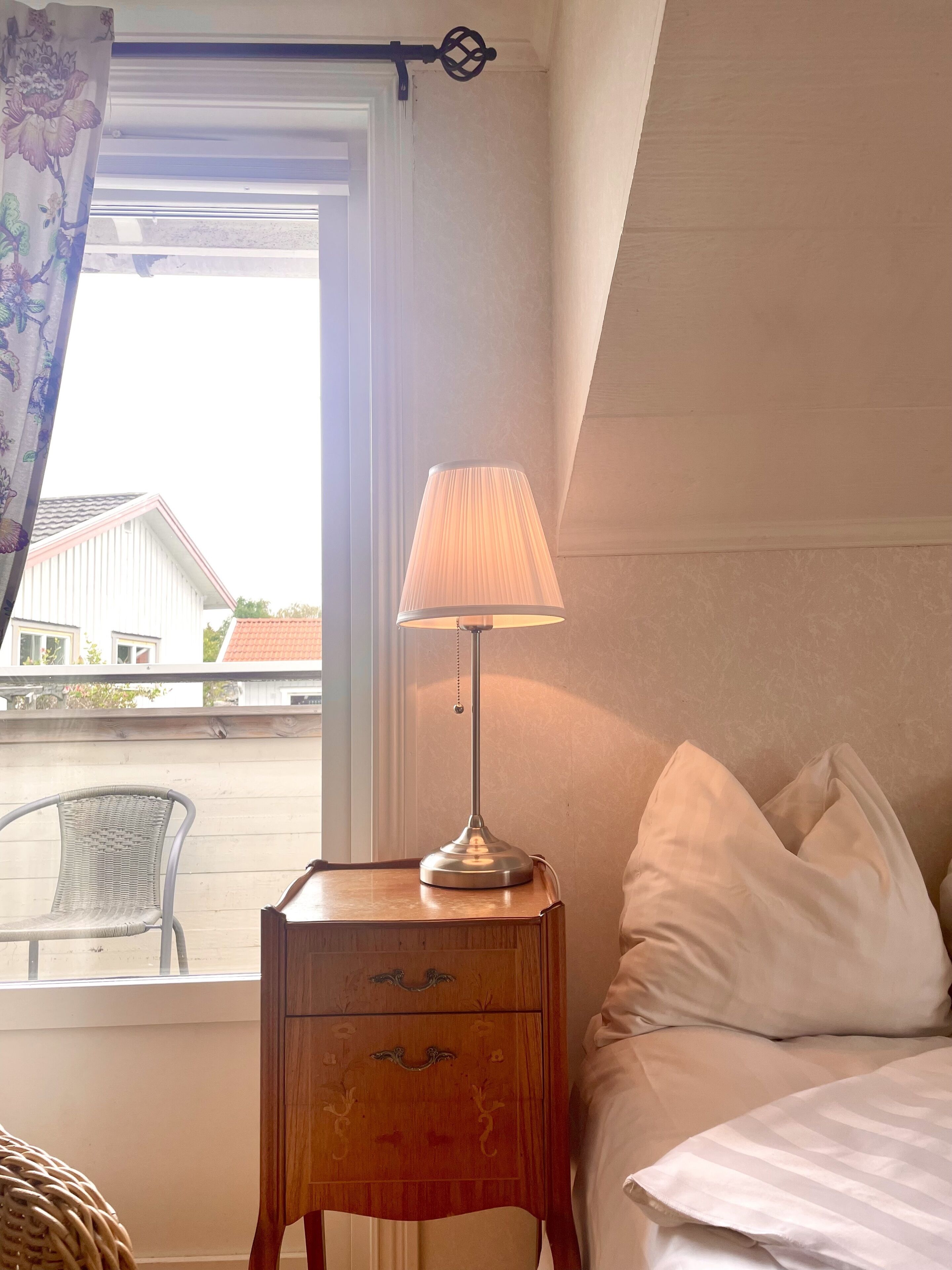 Una habitación con una cama, una mesita de noche con lámpara, una ventana con cortina y vistas a edificios.