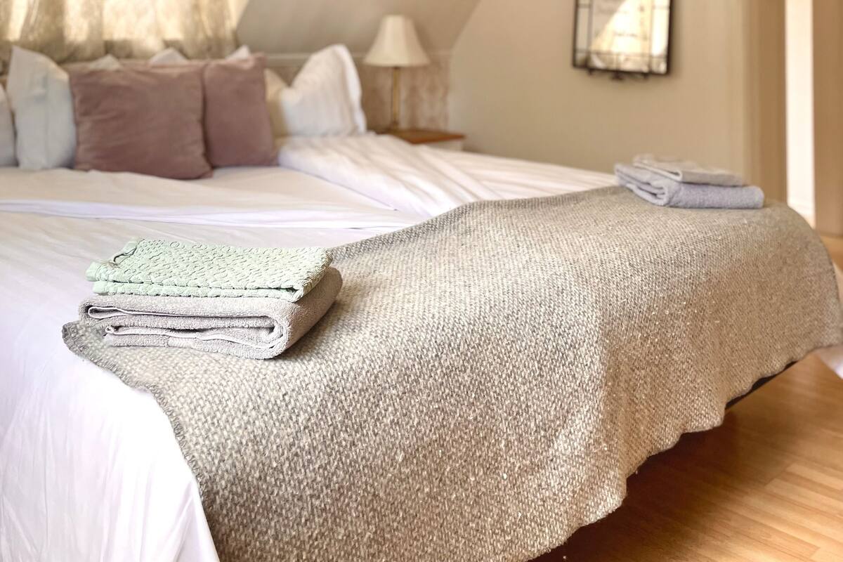 Une chambre avec un plafond en pente, un lit recouvert d’une couverture beige, des oreillers et une pile de serviettes pliées. On y trouve également un luminaire suspendu et un miroir au mur.
