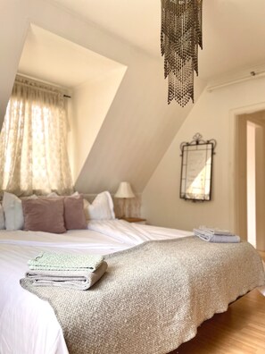 Een slaapkamer met een schuine kap, een bed met een beige deken, kussens en een stapel opgevouwen handdoeken. Er hangt een lamp en er is een spiegel aan de muur.