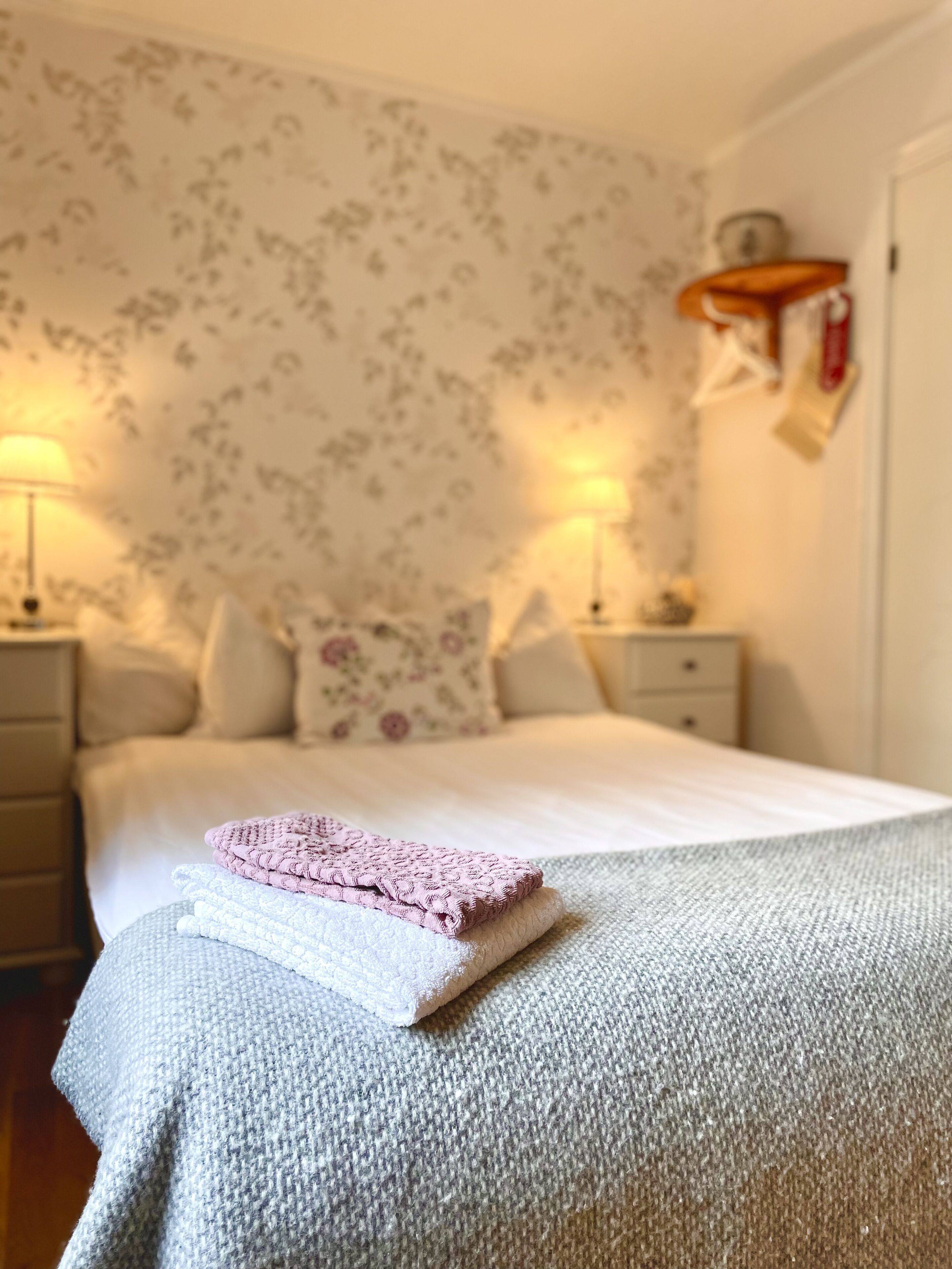 A neatly made bed with a pink and white blanket, a floral wallpaper, and two bedside lamps.