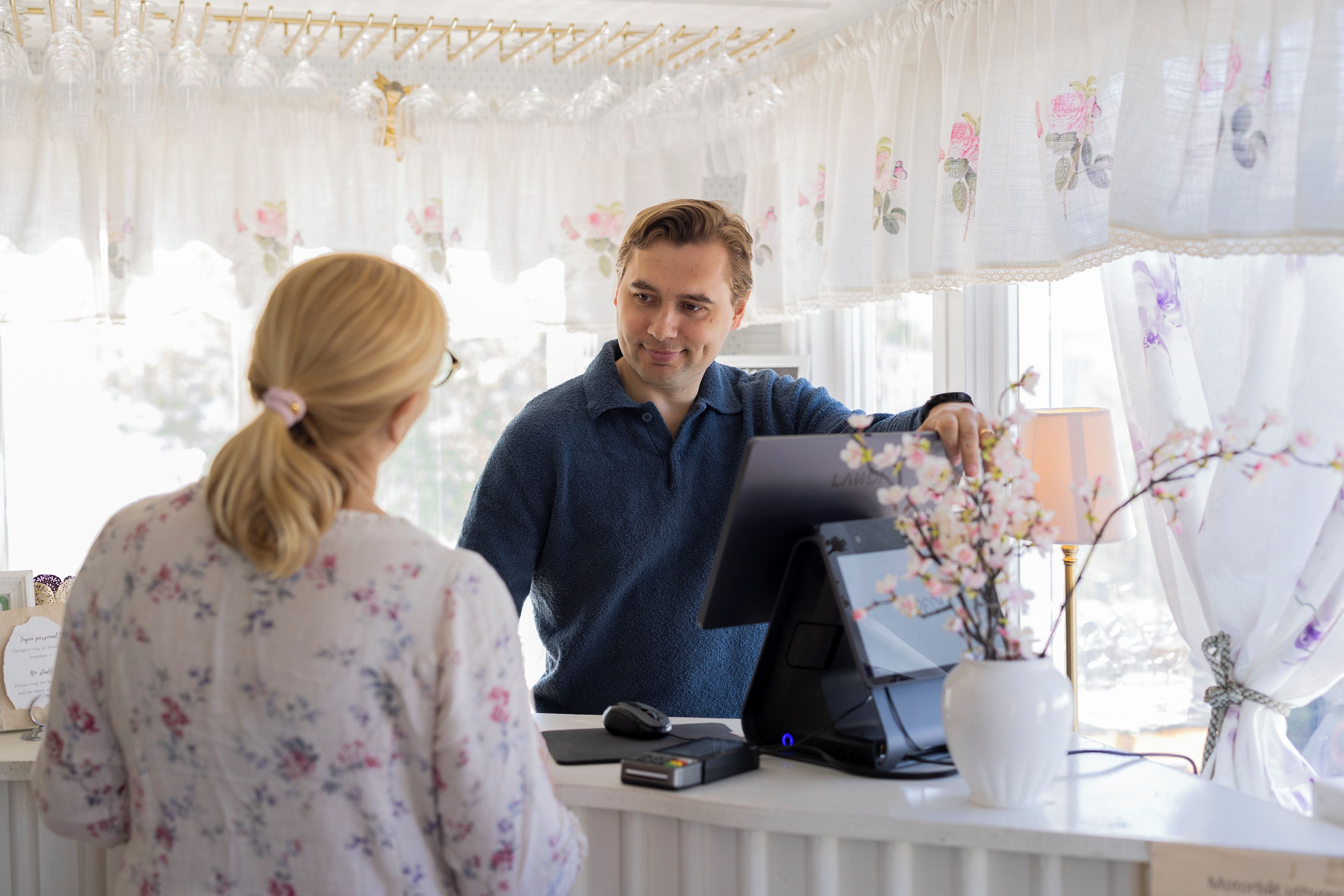 A person standing at a reception desk with a computer monitor, a mouse, and a keyboard. There is a vase with flowers on the desk.