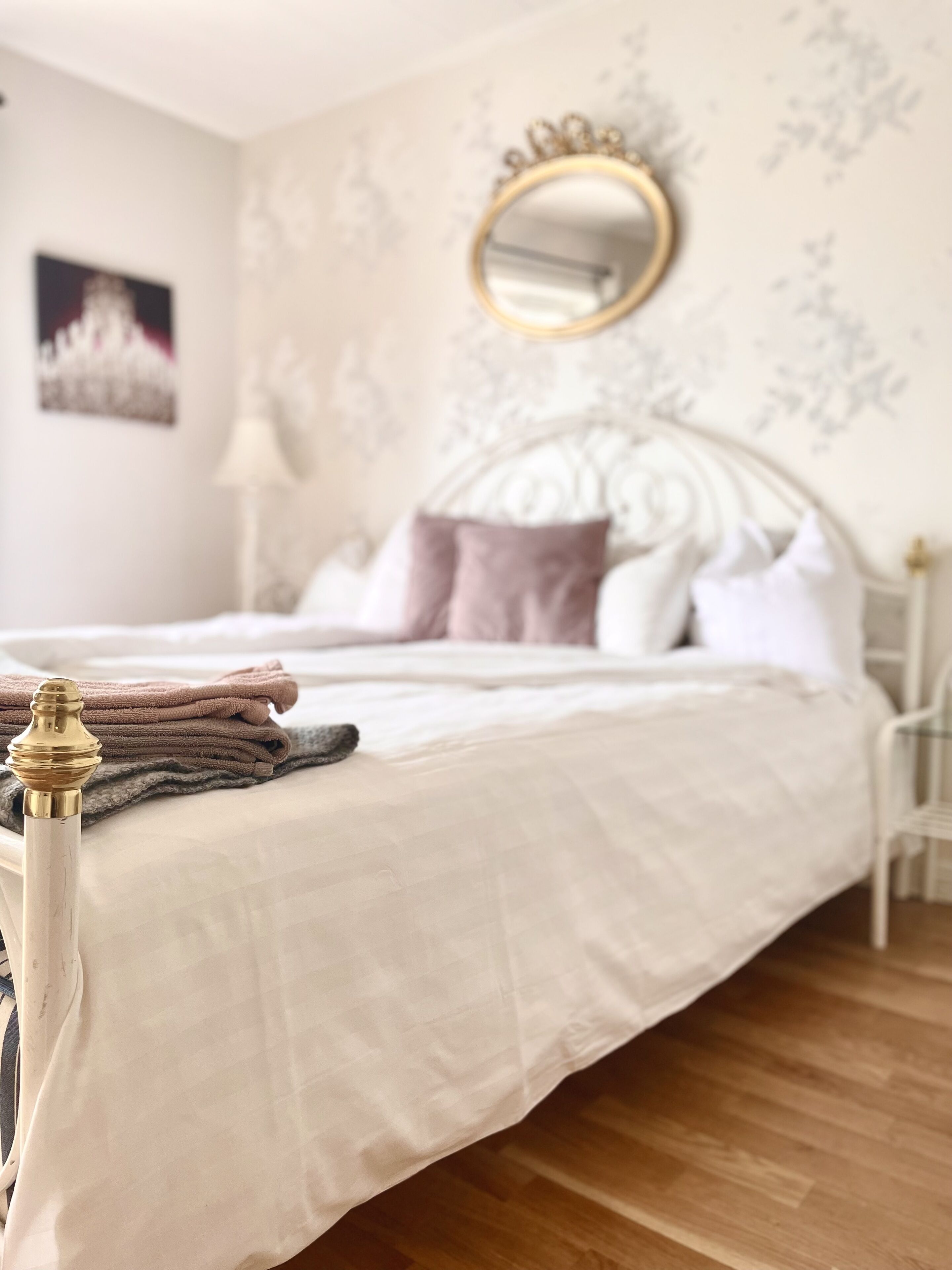 A neatly made bed with a white comforter and pillows, a gold-framed mirror on the wall, and a wooden floor.