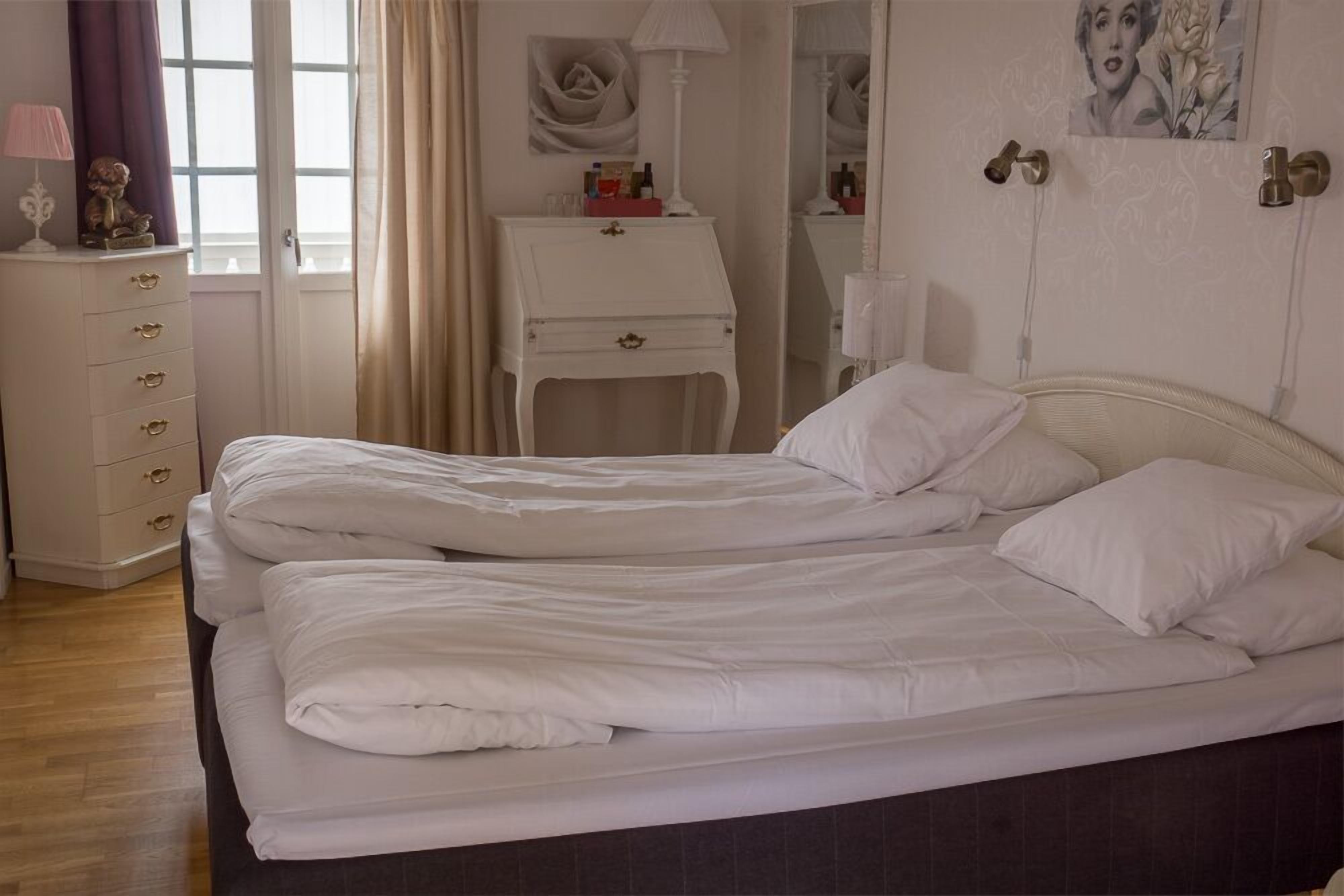 A double bed with white bedding, a white dresser with drawers, a lamp, and a mirror on the wall.