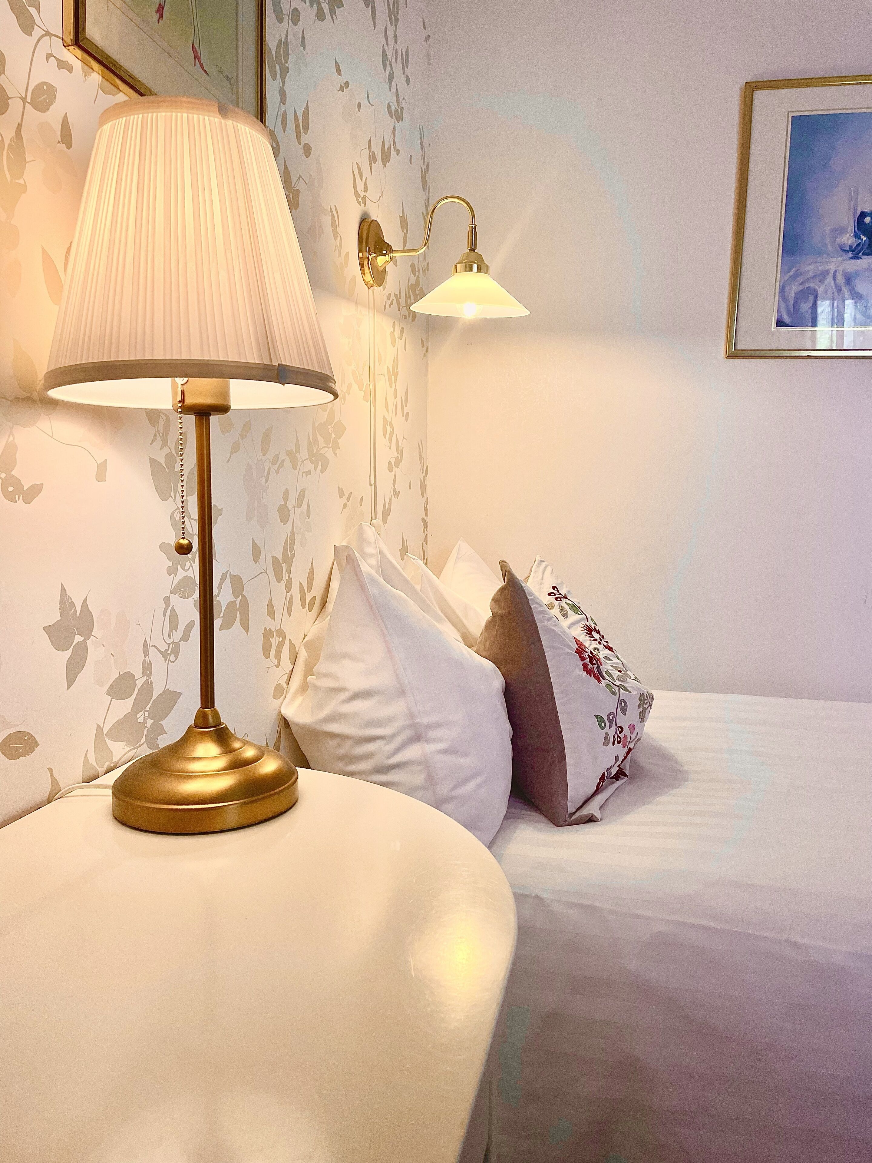 Una camera d'albergo con un letto, un comodino con una lampada e un punto luce a parete.