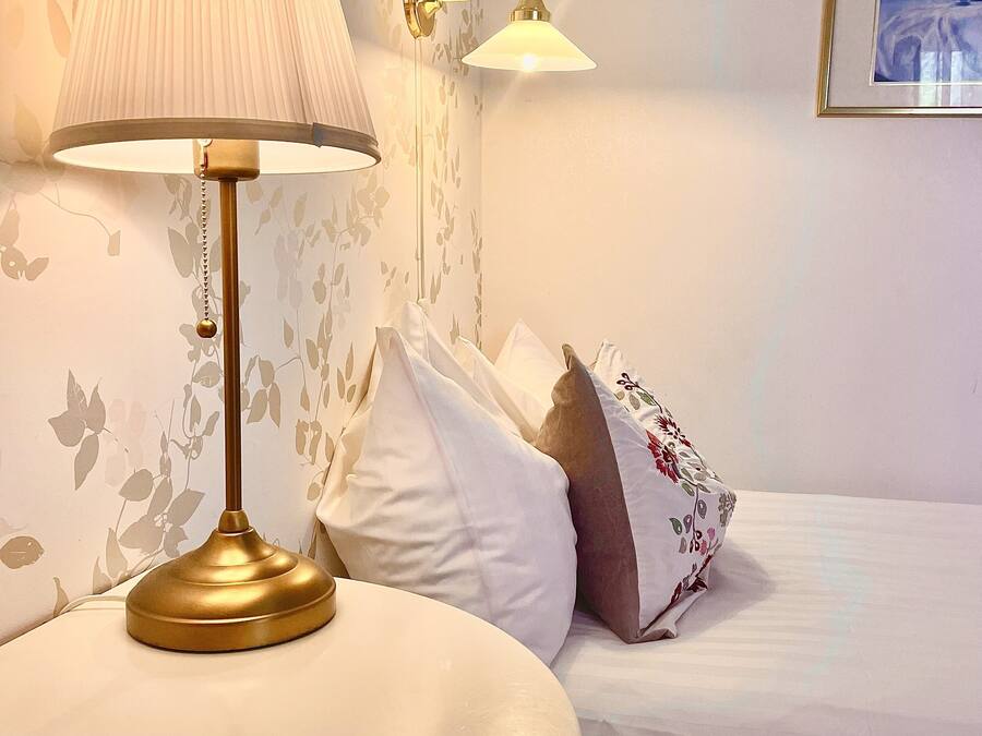 Una camera d'albergo con un letto, un comodino con una lampada e un punto luce a parete.