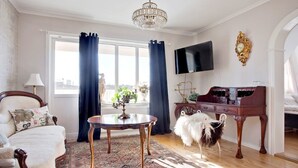 Une pièce avec une grande fenêtre, un lustre, une télévision, un piano et un chien blanc et duveteux.